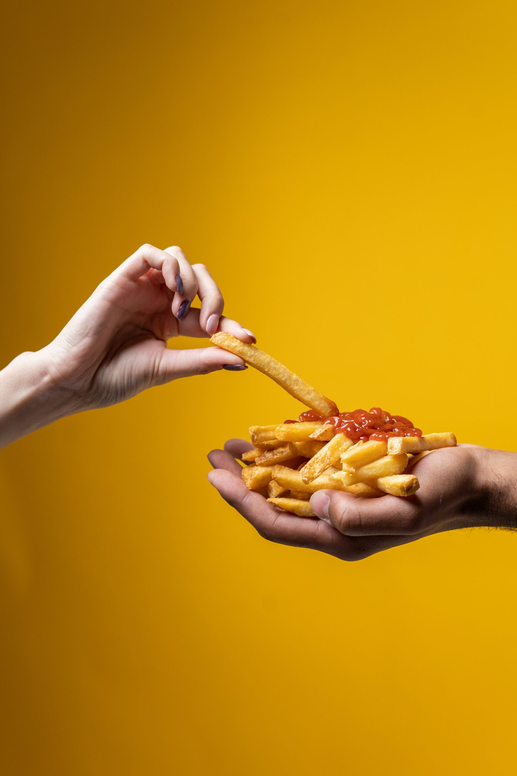 two outstretch hands hold a pile of french fries with some tomato sauce on the top. A third had reaches from the other side of the photo to dip a french fry in the sauce